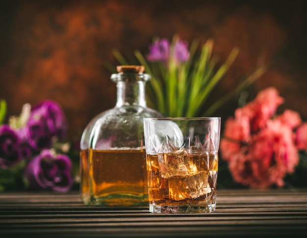 Vaso de whisky con hielo y botella con licor en una mesa de madera con algunas flores de color