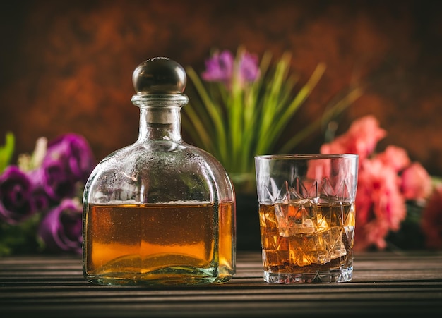 Foto vaso de whisky con hielo y botella con licor en una mesa de madera con algunas flores de color