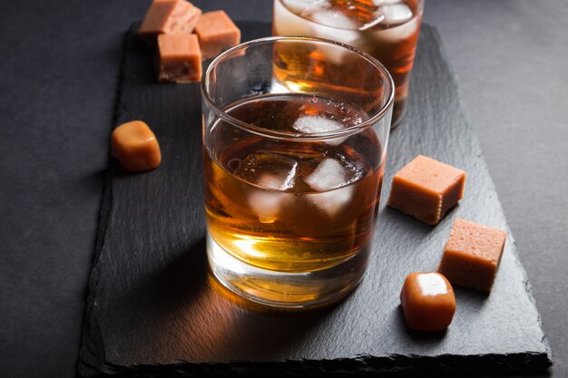 Vaso de whisky con hielo y caramelos de caramelo en un tablero de pizarra de piedra negra. Vista lateral