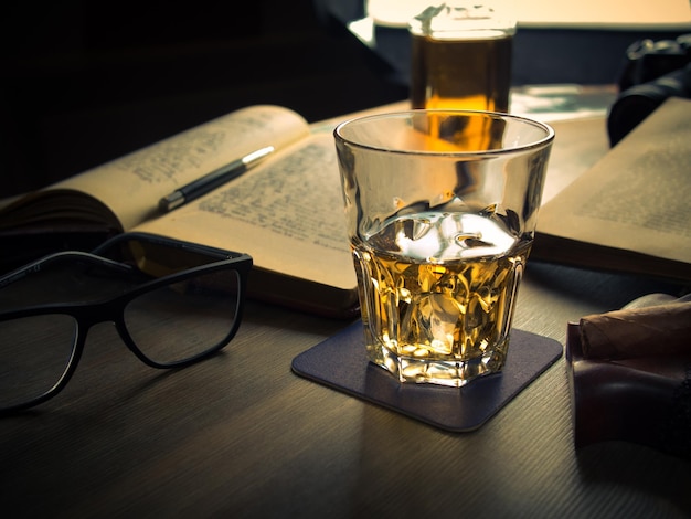 Vaso de whisky sobre una mesa con libros, cigarros y gafas.