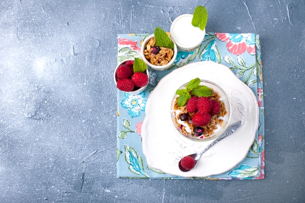 Un vaso con yogur, muesli y bayas sobre un fondo gris y un plato blanco