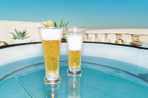 Vasos con cerveza fría con espuma en una mesa de cristal encima de la piscina en la terraza de un trópico