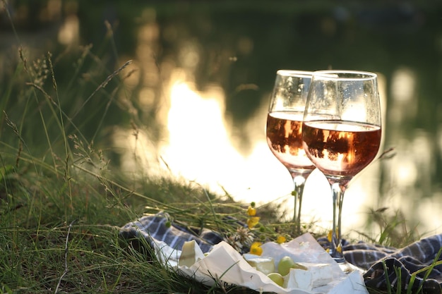 Vasos de delicioso queso de vino rosado y uvas en una manta de picnic cerca del espacio del lago para texto