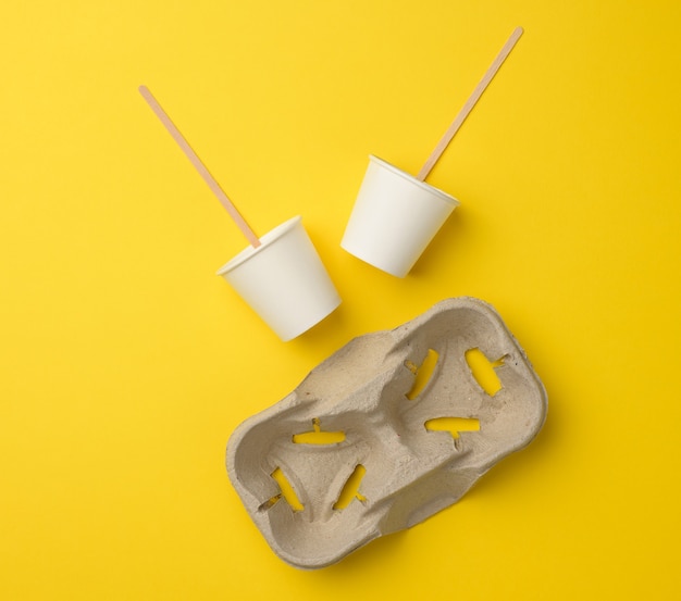 Vasos desechables de papel blanco, palos de madera y bandeja de papel sobre fondo amarillo. Envase de bebida para llevar
