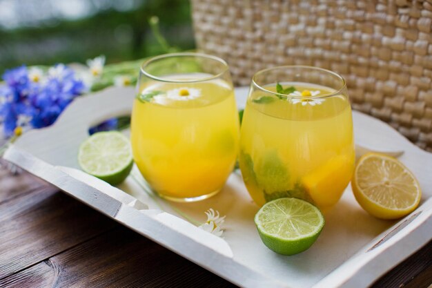 Vasos con limonada con menta, lima, limones y naranja en una bandeja de madera blanca cerca de una bolsa de paja con flores silvestres Concepto de bebidas de verano