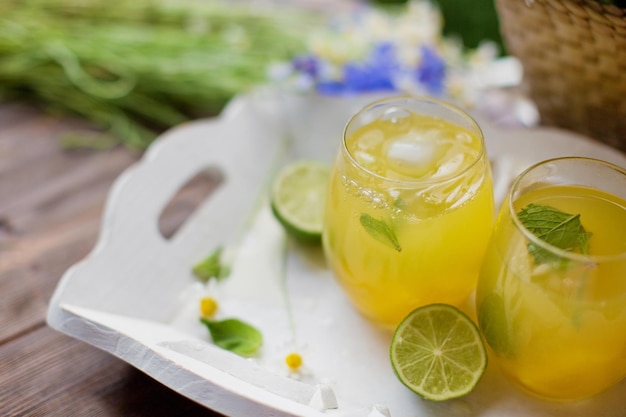 Vasos con limonada con menta, lima, limones y naranja en una bandeja de madera blanca cerca de una bolsa de paja con flores silvestres Concepto de bebidas de verano