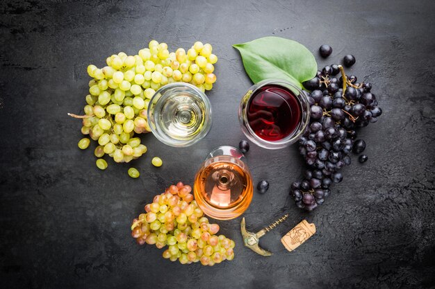 Vasos con vino blanco, tinto y rosado y uvas maduras sobre fondo de piedra negra, vista superior