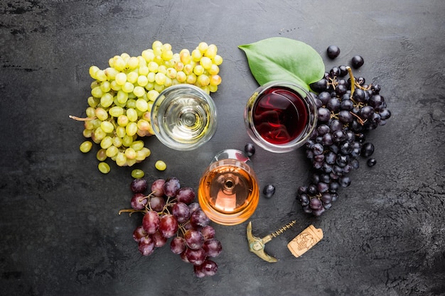 Vasos con vino blanco, tinto y rosado y uvas maduras sobre fondo de piedra negra, vista superior