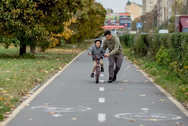 Vater bringt seinem Sohn das Fahrradfahren auf dem Radweg im Park bei. Der Vater hält ein Fahrrad und der Sohn sitzt darauf