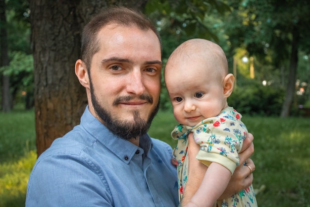 Vater, der Babysohn auf dem Hintergrund einer Sommerlandschaftsnahaufnahme hält. Bärtiger junger Mann, der lächelt, nachdenkliches Baby hält und in die Kamera schaut