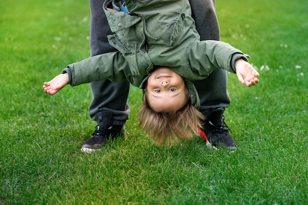 Vater, der mit kleinem Sohn draußen auf einem Gras spielt. Kleinkindjunge, der kopfüber schwingt. Glückliches Kind in einem Park. Aktives Elternkonzept.