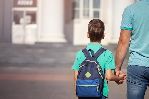 Vater führt ein kleines Schulkind von Hand in Hand. Elternteil und Sohn mit Rucksack hinter dem Rücken.