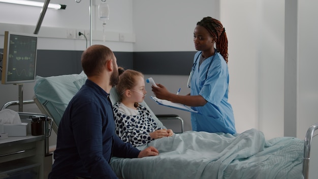 Foto vater hält kranke tochterhände während der ärztlichen untersuchung im krankenhauszimmer, die nach einer krankheitsoperation im bett ruht. afroamerikanische krankenschwester bringt infrarot-thermometer zur temperaturkontrolle