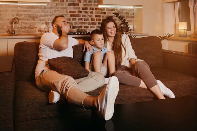 Vater mit Bart, Sohn und junge Mutter mit langen Haaren sehen auf dem Sofa fern