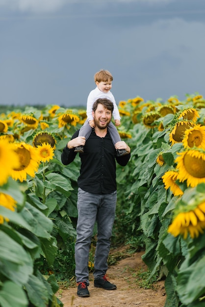 Vater mit einem kleinen Sohn auf seinen Schultern, der auf einem Sonnenblumenfeld spazieren geht und Spaß hat