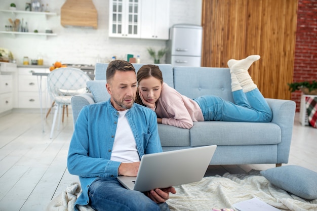 Vater mit einem Laptop sitzt auf dem Boden und Tochter liegt zu Hause auf dem Sofa, beide schauen auf den Laptop-Bildschirm und sind glücklich.