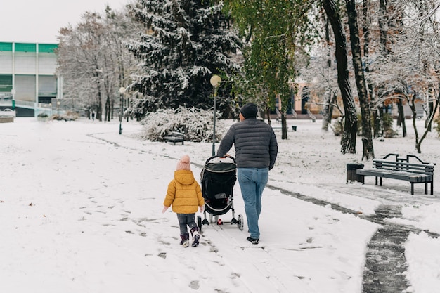 Vater mit Kind und Baby, die mit Kinderwagen im Winterschneepark gehen.