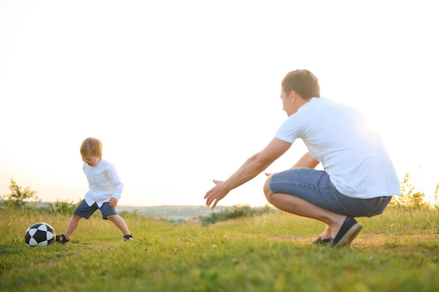 Vater mit kleinem Sohn spielt Fußball auf dem grünen Rasen im Park. Glückliche Familie, die Spaß hat und an einem sonnigen Tag auf einem grünen Rasen Fußball spielt. Familienkonzept: Vatertag