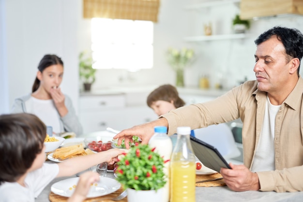 Vater mittleren Alters, der zusammen mit seiner Familie das Essen genießt und ein digitales Tablet verwendet, während er zu Hause am Tisch in der Küche sitzt. Glückliche Familie, Technologie, Morgenkonzept