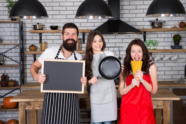 Vater Mutter und Kind kochen Mutter Vater mit kleinem Mädchen Familientag Glückliche Familie in der Küche Kleines Mädchen mit den Eltern in Schürze Bärtiger Mann mit Brettkopierraum Kochen mit Leidenschaft