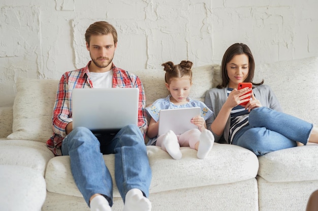 Vater, Mutter und Tochter mit elektronischen Geräten sitzen auf dem Sofa im Wohnzimmer.