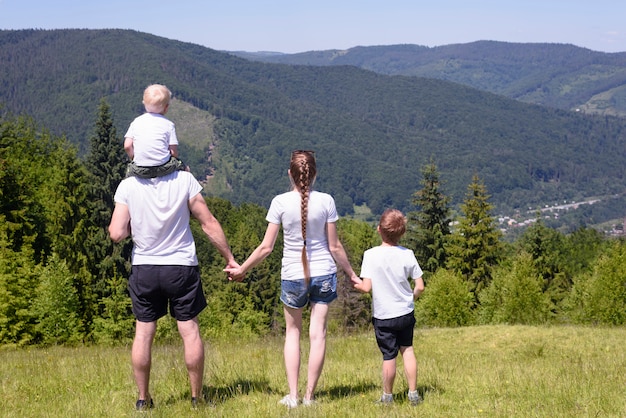 Vater, Mutter und zwei kleine Söhne stehen auf einer grünen Wiese bewaldeter Hügel. Familienwerte