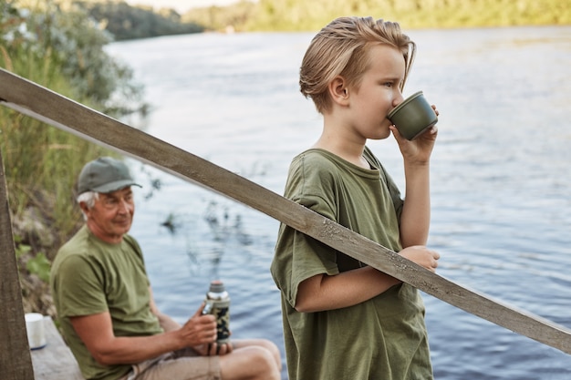 Vater sitzt auf Holzplatz mit Thermoskanne in den Händen und schaut in die Ferne zum Fluss, sein Sohn trinkt heißen Tee und genießt die schöne Natur.