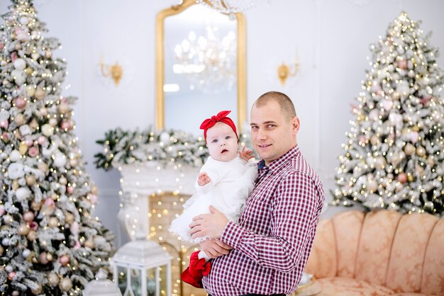 Vater spielt mit einem Mädchen in einem weißen Kleid, umarmt sie, küsst sie. Der Hintergrund des festlichen Weihnachtsbaumes