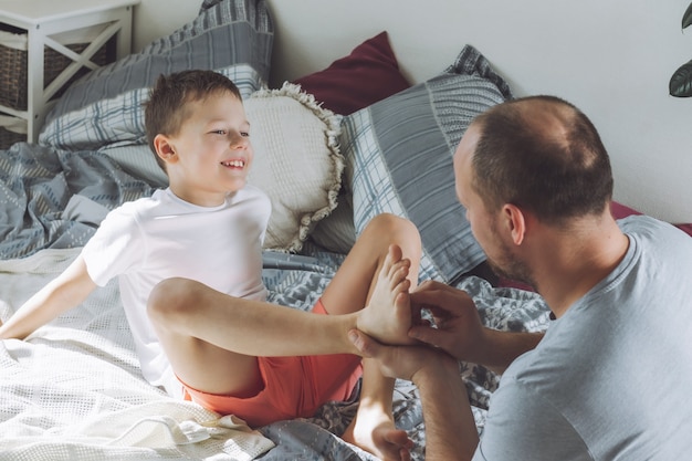 Vater spielt mit seinem Sohn 7-10 im Bett. Papa kitzelt Kinderfüße. Familie, viel Spaß