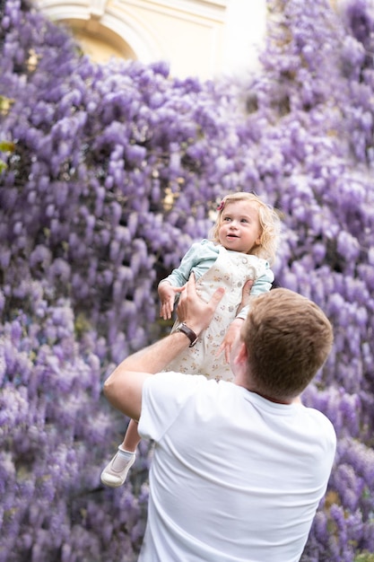 Vater spielt Spaß im Freien mit Baby-Mädchen-Tochter auf dem Hintergrund der Glucinum-Blume lila sehr peri