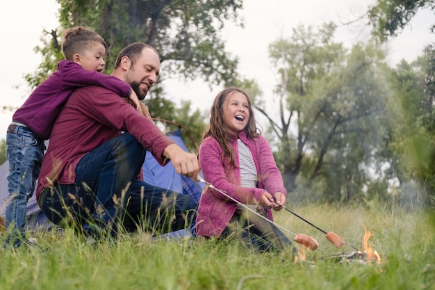 Vater, Tochter und Sohn sitzen am Lagerfeuer im Wald und braten Würstchen. Freizeit mit Vater, Elternschaft. Glückliches Familienkonzept