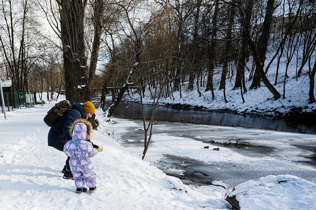 Vater und Kind füttern an einem sonnigen, frostigen Wintertag im Park Enten auf dem zugefrorenen Fluss