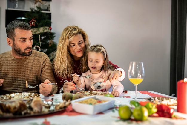 Vater und Mutter sitzen mit ihrer Tochter am Tisch und feiern das Weihnachtsessen Mama füttert ihr kleines Mädchen