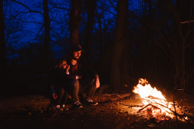 Vater und sein kleiner Sohn sitzen nachts zusammen auf den Baumstämmen vor einem Feuer. Die Wanderung im Wald. .