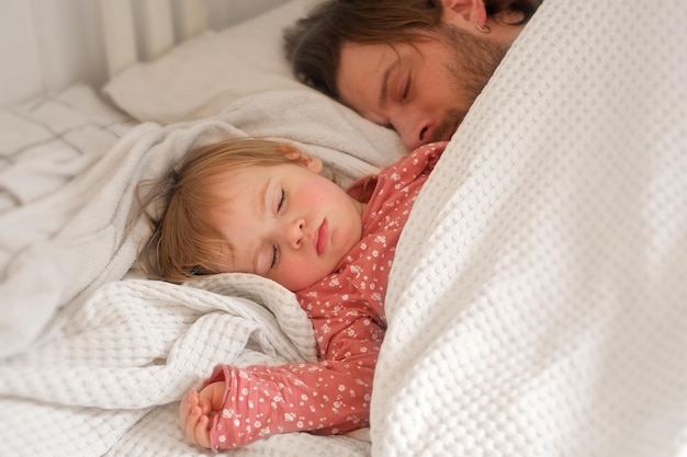 Vater und seine kleine Tochter schlafen zusammen. Gesunder Schlaf. Glückliche liebevolle Familie. Eltern und Kind schlafen zu Hause im weißen Bett. Authentischer Lebensstil. Vaterschaftsmomente. Das Leben eines alleinerziehenden Vaters