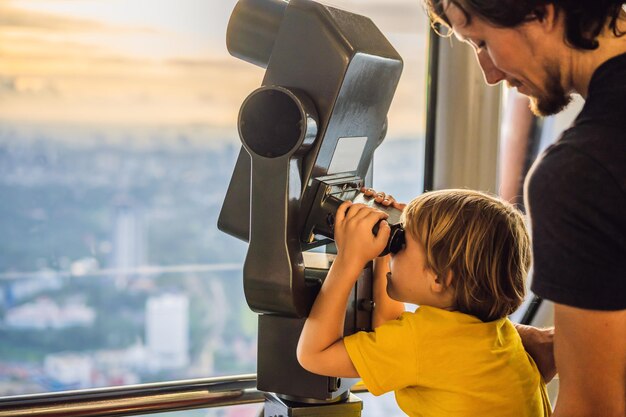 Vater und Sohn betrachten das Stadtbild von Kuala Lumpur Verwenden Sie ein Fernglas Panoramablick auf die Skyline von Kuala Lumpur Abend bei Sonnenuntergang Wolkenkratzer Gebäude in Malaysia Reisen mit Kindern Konzept