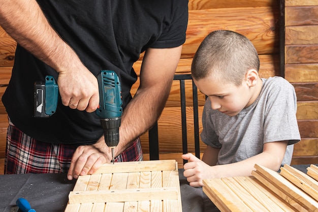 Vater und Sohn bohren in der Werkstatt mit einem Schraubenzieher, Werkzeugen und einem Balken ein Holzbrett auf den Tisch