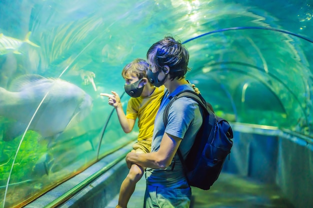 Vater und Sohn in medizinischen Masken schauen sich die Fische im Aquarium im Ozeanarium an. Menschen gehen medizinisch