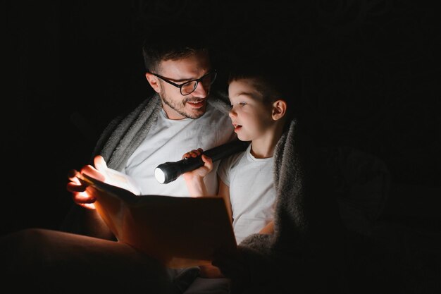 Vater und Sohn mit Taschenlampe lesen Buch unter der Decke zu Hause