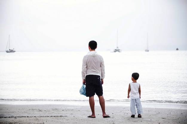 Vater und Sohn schauen auf das Meer Stehen Sie zurück mit Blick auf das Wasser Sandstrand im Hintergrund der Yacht