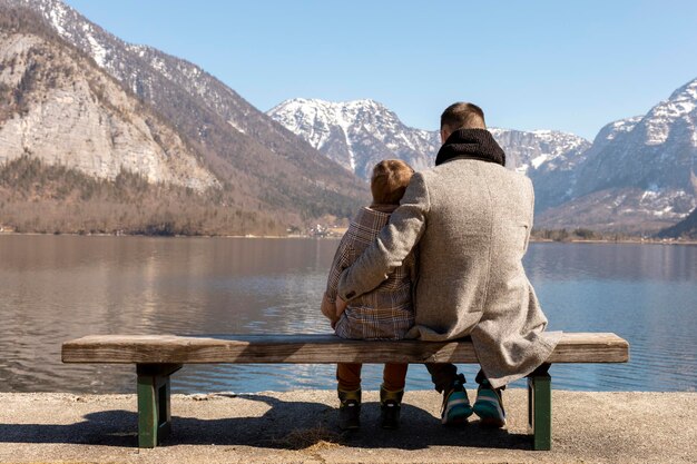 Vater und Sohn sitzen zusammen draußen auf der Bank und genießen Berge Schnee gutes Wetter blauer Himmel Kleiner Junge und sein Vater verbringen Zeit zusammen Zeit mit der Familie Schöne Winterlandschaft