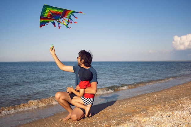 Vater und Sohn stehen an einem Sandstrand am Meer und starten im Sommer im Urlaub einen gestreiften Spielzeugdrachen