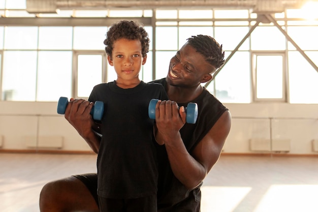 Vater und Sohn treiben im Fitnessstudio Sport. Ein Afroamerikaner hilft seinem Sohn beim Hantelheben