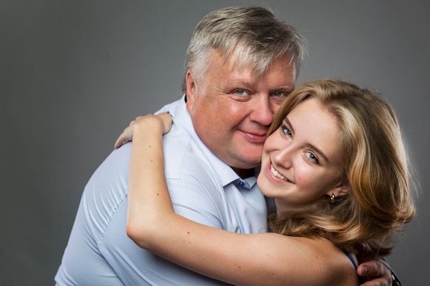 Vater und Tochter im Teenageralter lachen und umarmen Ein dicker Mann in einem Hemd und ein schönes Mädchen in einem blauen Kleid auf dunkelgrauem Hintergrund Liebe und Zärtlichkeit Nahaufnahme
