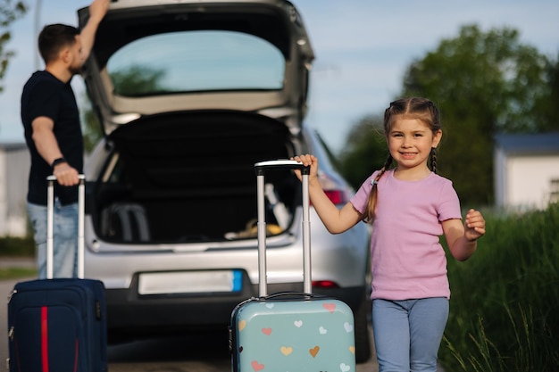 Vater und Tochter stehen mit ihren Koffern in der Nähe eines Autos mit einem offenen Kofferraum und einem glücklichen kleinen Mädchen