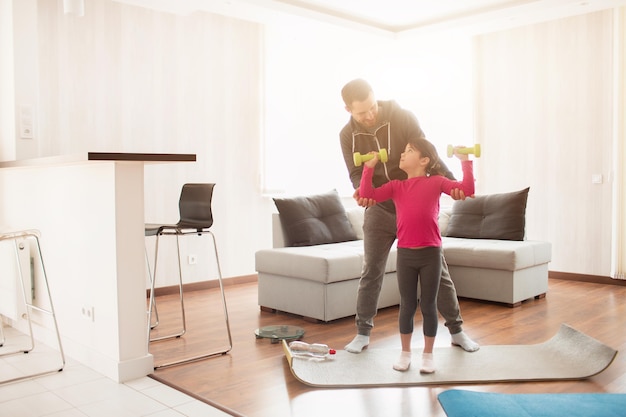 Vater und Tochter trainieren zu Hause. Training in der Wohnung. Sport zu Hause. Sie stehen auf einer Yogamatte. Mädchen, das Hantel hält.