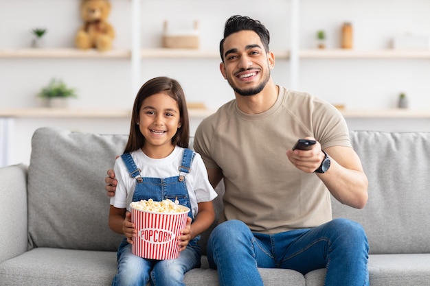 Vatertag. Porträt eines glücklichen jungen Mannes und eines Mädchens, die Fernsehsendungen oder Filme ansehen, zu Hause auf der Couch im Wohnzimmer sitzen und Spaß haben. Aufgeregter Vater mit Fernbedienung, Tochter isst Popcorn