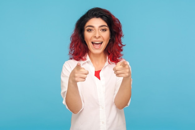 Vaya, oye, tienes suerte. Retrato de mujer hipster feliz sorprendida con el pelo rojo en camisa señalando con el dedo a la cámara y expresando asombro, eligiendo al ganador. Foto de estudio interior aislado sobre fondo azul.