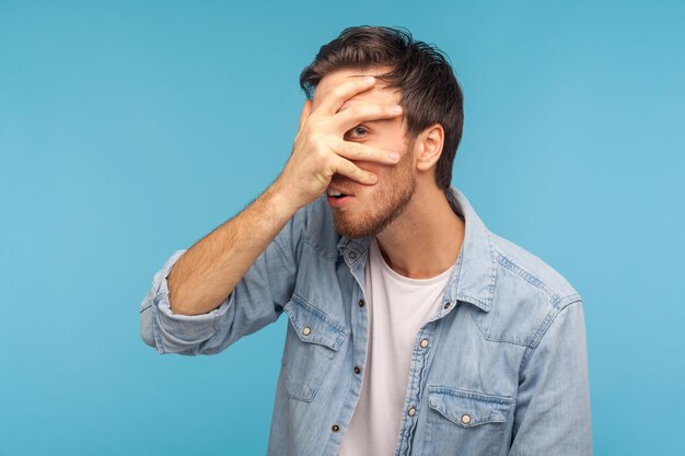 Foto vaya, qué es. retrato de un curioso hombre entrometido con pantalones de denim trabajador mirando a través de los dedos con expresión sorprendida e inquisitiva, viendo contenido prohibido, espiando secretos. tiro del estudio aislado