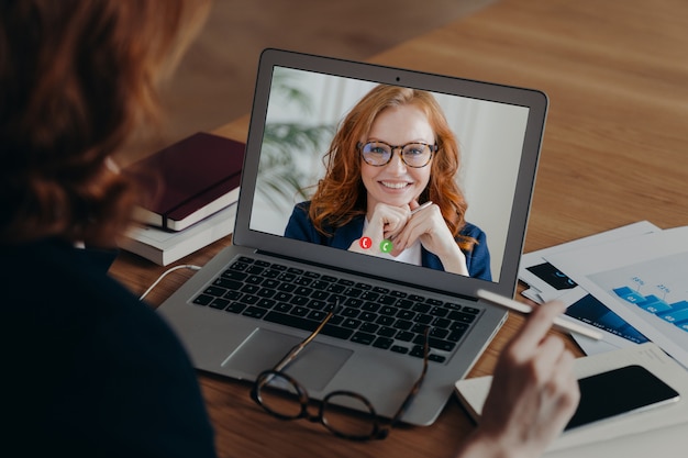 Vea por encima del hombro de las mujeres autónomas conversaciones con los empleados por video llamada, discuta la estrategia de trabajo. Opinión de la pantalla del ordenador portátil de la mujer sonriente del jengibre en gafas. Uso de nueva tecnología. Trabajo a distancia